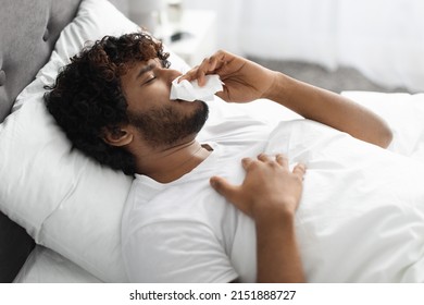 Seasonal Virus, Coronavirus, Flu, Allergy Concept. Closeup Photo Of Sick Indian Millennial Guy Blowing His Nose, Lying In Bed With Fever, Side View, Copy Space, Home Interior