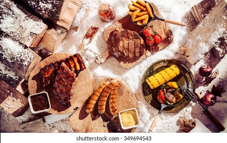 Seasonal winter barbecue in snow with grilled spicy steak, ribs and sausages served with assorted grilled vegetables and potato wedges, overhead view