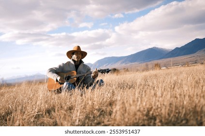 A seasoned cowboy sits in a field of dry grass and plays the guitar. A single free man in a cowboy hat holds a guitar in his hands. The sky is overcast, mountains and hills rise behind. - Powered by Shutterstock