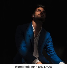 Seated Elegant Man Looks Up With Ray Of Light On His Face On Dark Background