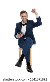 Seated Gentleman In Blue Tux Celebrating Victory While Holding A Mobile Phone On White Background