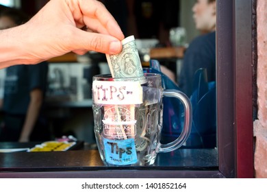 Seattle WA May 18 2019. A tips mug standing on the cafe counter, a hand  of a tipping customer is adding a dollar bill to it. Focus on the mug.