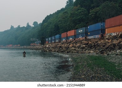 Seattle, WA, USA - August 20, 2018: BNSF Freight Train Traveling Through Carkeek Park During A Smoky Evening With Single Man Fishing In Puget Sound