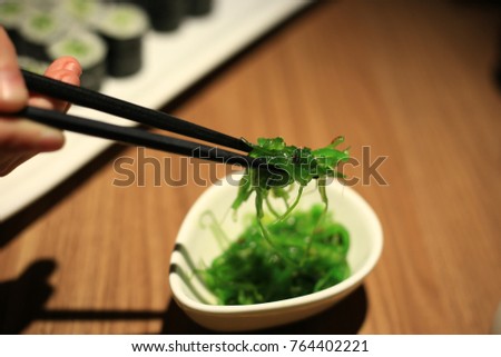 Seaweed salad lit dramatically on a white dish on a wooden table with a womans hand and chopsticks.