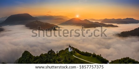 Sebrelje, Slovenia - Aerial panoramic drone view of the beautiful hilltop church of St.Ivan (Sv. Ivan Cerkev) at sunrise with huge morning fog bellow the valley and Julian Alps at background