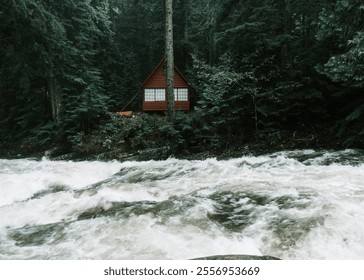 A secluded red cabin nestled among dense evergreen trees, with a rushing river in the foreground creating a peaceful and moody Pacific Northwest scene - Powered by Shutterstock