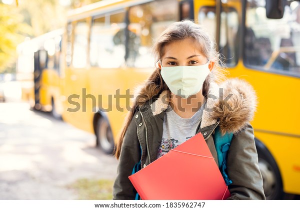 Secondary school girl learner in protective mask\
in front of school bus, ready to go home by bus. School education\
during the covid-19\
pandemic