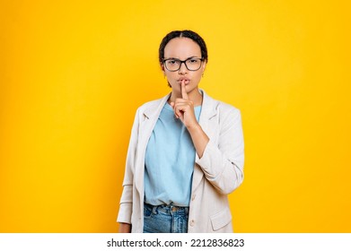 Secret Concept.Pretty, Cautious Hispanic Or Brazilian Young Business Woman With Glasses, Holds Finger Near Lips, Saying Hush Be Quiet, Shhh Gesture, Stand On Isolated Orange Background,looks At Camera