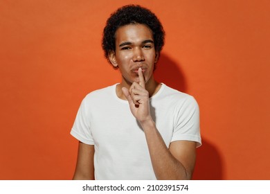 Secret Fun Young Black Curly Man 20s Years Old Wears White T-shirt Say Hush Be Quiet With Finger On Lips Shhh Gesture Lookng Camera Blinking Isolated On Plain Pastel Orange Background Studio Portrait