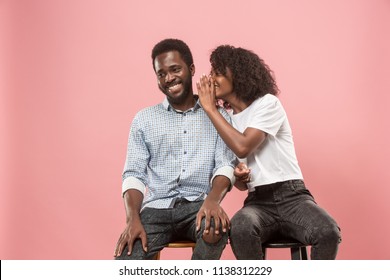 Secret, gossip concept. Young woman whispering a secret behind her hand to afro man. African couple isolated on trendy pink studio background. Young emotional afro woman and man. Human emotions