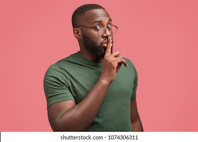 Secret man with dark skin, makes silence gesture, says: Hush, stop talking, looks seriously at camera, wears round spectacles and dark green t shirt isolated over pink background. African guy gestures