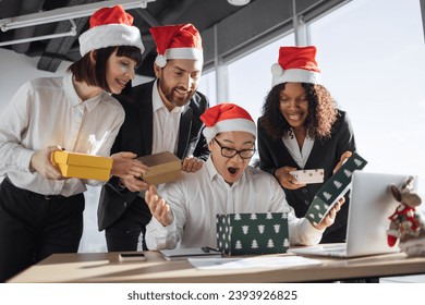 Secret Santa. Smiling Asian businessman in Santa hat unpacks a Christmas present from multinationals colleagues at work standing around him. New Year's eve. - Powered by Shutterstock