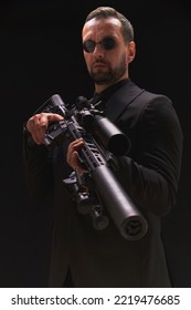 Secret Service Man In Black Suit, Bodyguard Or Special Forces Agent Man With Gun Isolated On Black Background. Man In Black Suit Holding Assault Rifle. Spy, Police Man In Black Suit And Sunglasses.