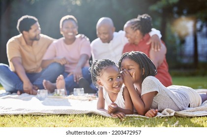 Secret, sister and children with a girl whispering to her sibling and a black family in the background. Kids, mystery and gossip with a female child being secretive on a picnic in the park in summer - Powered by Shutterstock