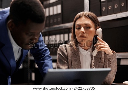 Secretary talking with bookkeeper at landline phone, working together at accountancy report in corporate depository. Diverse businesspeople analyzing administrative documents in file room