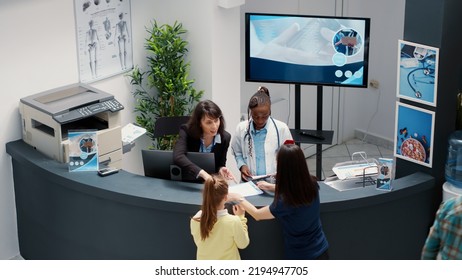 Secretary Working At Busy Hospital Reception Desk To Help Patients With Examination Appointment In Waiting Room. Receptionist Giving Medical Reports To Group Of People In Clinic Lobby. Handheld Shot