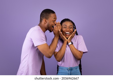 Secrets And Gossips Concept. Young Black Man Sharing News With His Excited Girlfriend, Whispering To Ear, Surprised Woman Raising Hands In Amazement Standing Together Over Purple Violet Background