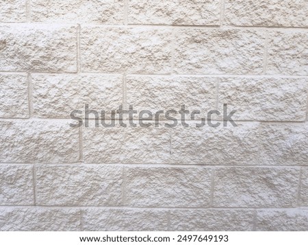 A section of a beige brick wall with a clean, smooth concrete sidewalk in the foreground. The wall textured surface and uniform brick pattern provide a neutral backdrop, while the sidewalk adds a