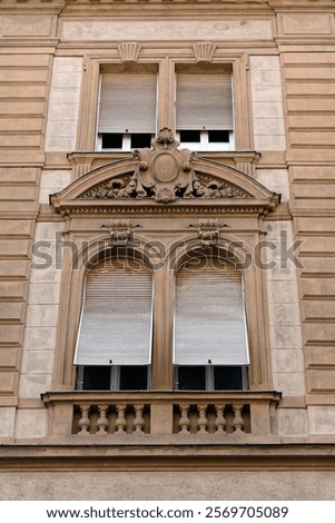 A section of a building facade in Budapest, featuring two sets of arched windows with closed blinds and an ornate decorative element above the lower windows. 