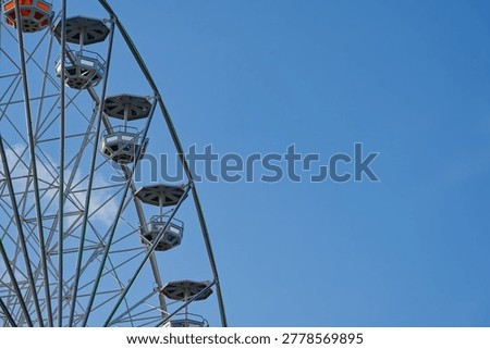 A section of a Ferris wheel with empty cabins is shown against a bright blue sky, highlighting the metal structure and colorful lights on a clear day.