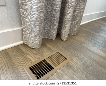 A section of gray patterned curtains hangs just above a floor vent on wood laminate flooring. The space is bright and minimal.