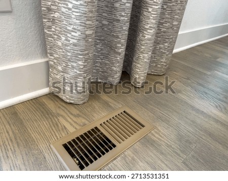 A section of gray patterned curtains hangs just above a floor vent on wood laminate flooring. The space is bright and minimal.