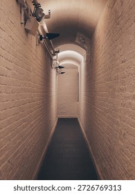 Secure Brick Corridor with Vaulted Ceiling and Surveillance Cameras. A narrow corridor with light-colored brick walls and a vaulted ceiling, illuminated by warm overhead lighting. Multiple black surveillance cameras are mounted along the left wall, emphasizing a sense of security and control. The dark tiled floor contrasts with the ambient lighting, creating a moody, symmetrical composition ideal for architectural or security-themed visuals.