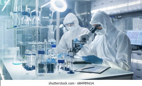 In a Secure High Level Laboratory Scientists in a Coverall Conducting a Research. Chemist Adjusts Samples in a  Petri Dish with Pincers and then Examines Them Under Microscope. - Shutterstock ID 684989614