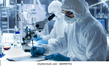 In A Secure High Level Laboratory Scientists In A Coverall Conducting A Research. Chemist With His Colleague Writes Down Experiment Results.