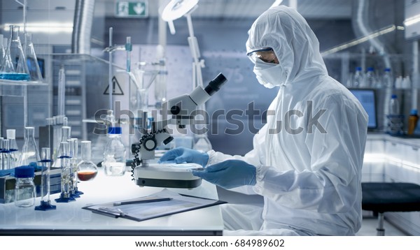 In a Secure High\
Level Laboratory Scientists in a Coverall Conducting a Research.\
Biologist Adjusts Samples in a Petri Dish with Pincers and Examines\
Them Under Microscope.