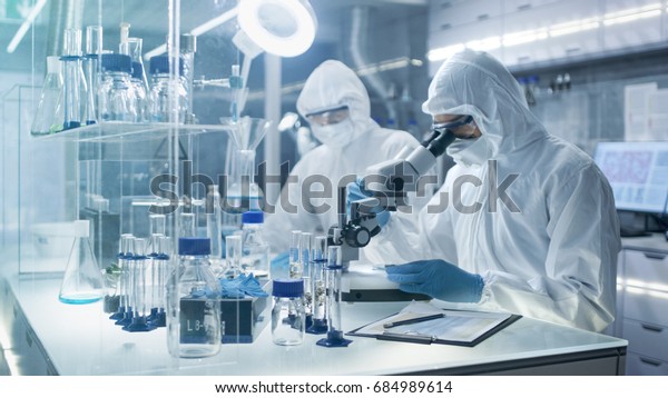 In a Secure High\
Level Laboratory Scientists in a Coverall Conducting a Research.\
Chemist Adjusts Samples in a  Petri Dish with Pincers and then\
Examines Them Under\
Microscope.