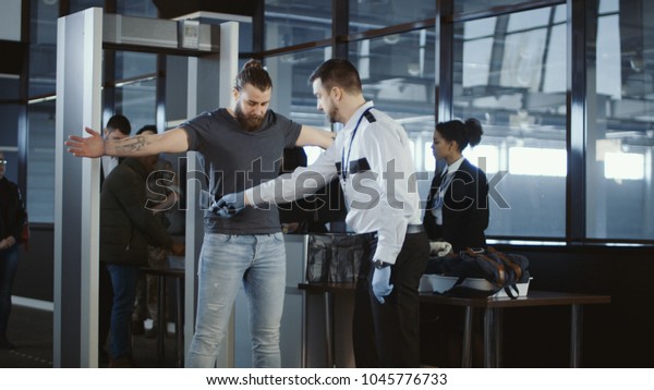 Security\
agent at an airport check-in gate patting down a bearded casual\
male passenger with outstretched arms after he passes through the\
metal detector scanner in the departures\
hall.