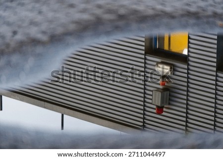 A security alarm unit appears reflected in a puddle in front of a metal clad industrial facade. Strong lines, soft focus edges make this scene ideal for safety, surveillance and infrastructure themes