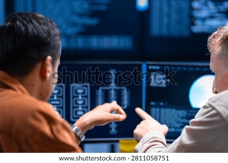A security analyst shows a finding on the monitor to a colleague at a multi-monitor workstation in a darkened control room. The frame illustrates defending the corporate network.
