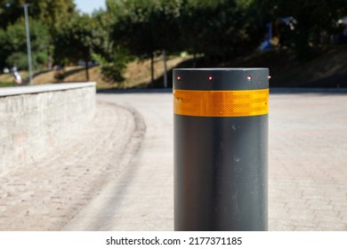 Security Bollard To Block Or Enable Traffic. Bollard With Warning Light For Physical And Visual Barrier. Black Steel Bollard For Road Traffic Control. Metallic Bollard With Orange Safety Reflector