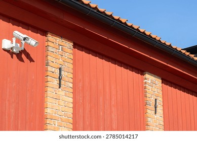 Security camera attached to a red barn wall with brick columns, under a clear blue sky, highlighting contemporary surveillance features