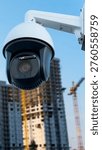 A security camera is fixed on a construction site where workers are visible in the background. The camera captures real-time actions in a busy area. Scaffolding and cranes are present.
