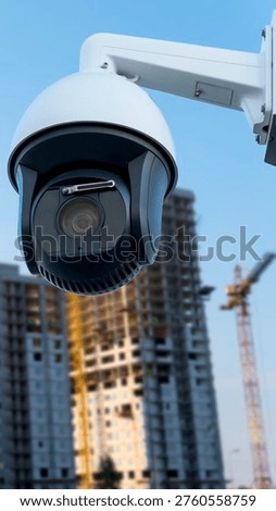 A security camera is fixed on a construction site where workers are visible in the background. The camera captures real-time actions in a busy area. Scaffolding and cranes are present.