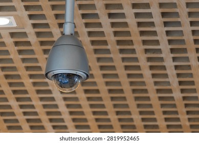 Security camera hangs from wooden grid ceiling. Dome-style surveillance device, gray and round. Used in public spaces, offices, or retail stores. Monitors movement, ensures safety