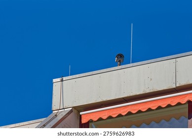 A security camera installed on the rooftop of a building under a bright blue sky, featuring a red awning and concrete structure.