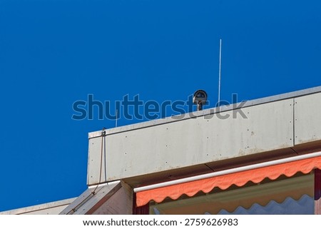 A security camera installed on the rooftop of a building under a bright blue sky, featuring a red awning and concrete structure.