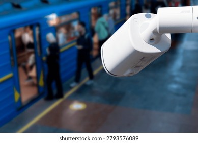 A security camera looks down at a subway station. People wait by the blue train. The scene shows passengers getting on and off as the train arrives during evening hours.