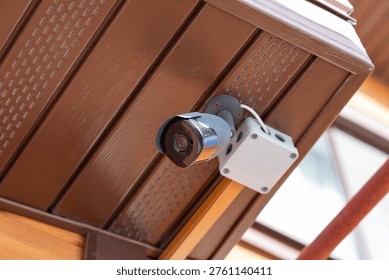 Security camera mounted on brown ceiling under roof. Surveillance system for property protection and safety monitoring. Digital video equipment for prevention of burglary and crime control.