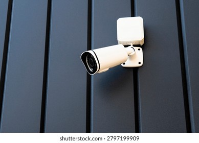 A security camera is mounted on a dark wall. It is positioned to monitor the area. The camera is ready to capture any movement. This setup helps keep the place secure during the day and night.