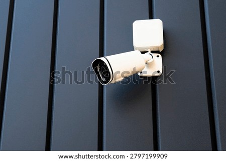 A security camera is mounted on a dark wall. It is positioned to monitor the area. The camera is ready to capture any movement. This setup helps keep the place secure during the day and night.