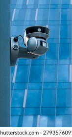 A security camera is mounted on a post, watching over a modern building with a glass facade. The building reflects the blue sky with clouds, showing urban life in progress.