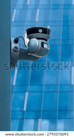 A security camera is mounted on a post, watching over a modern building with a glass facade. The building reflects the blue sky with clouds, showing urban life in progress.