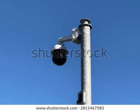 A security camera is positioned on a tall pole against a bright blue sky in a city.