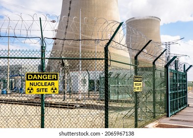 Security fence of a nuclear power station with yellow danger warning signs and barbed wire, and two cooling towers in the background. - Shutterstock ID 2065967138