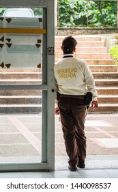 Security guard from the back standing at the door of a building. He's wearing a yellow shirt (it says private security, Spanish) and brown pants. He leans against the door. Taken in Armenia, Colombia